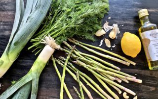 Fresh asparagus, leek, dill, garlic, lemon, and cashews on a dark wooden surface.