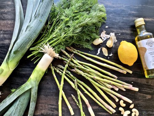 Fresh asparagus, leek, dill, garlic, lemon, and cashews on a dark wooden surface.