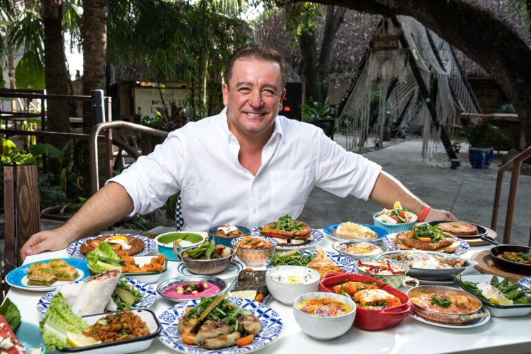 Smiling man in white shirt surrounded by diverse dishes at a table.