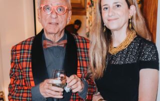 Two people posing at an indoor event, one in a red plaid jacket holding a drink.