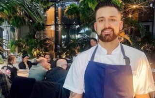 Chef in a blue apron smiling inside a lively restaurant with greenery.