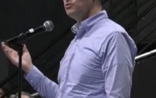 Man in a blue shirt speaking at a microphone in front of an audience.