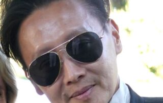 Man wearing aviator sunglasses and a suit, smiling slightly.