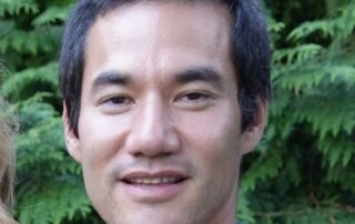 Close-up of a smiling man outdoors with green foliage in the background.