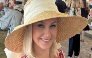 A smiling woman wearing a large straw hat and floral dress at an outdoor event.