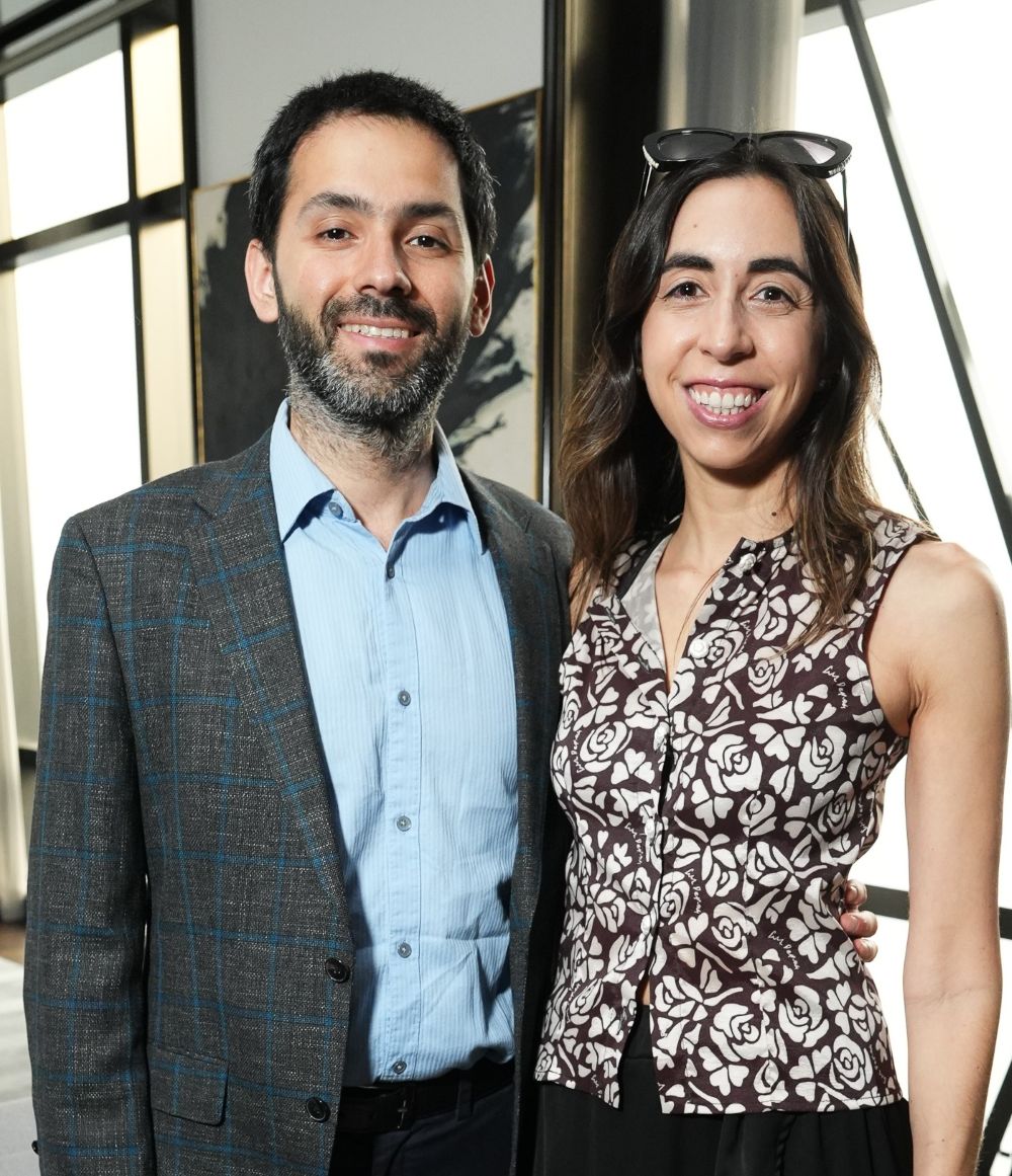 A smiling couple posing indoors near a window, dressed in smart casual attire.