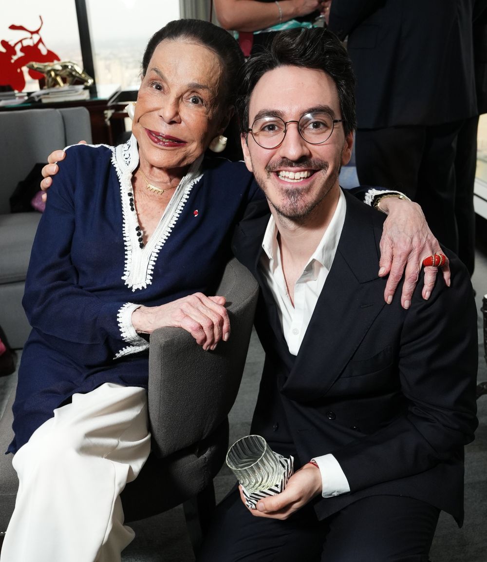 An elderly woman and a young man smile together in a warm embrace indoors.