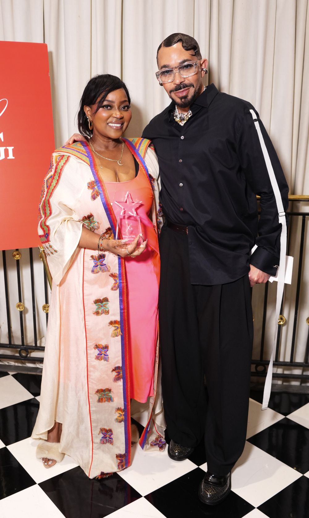 A woman in a colorful robe holding an award stands next to a man in black attire.