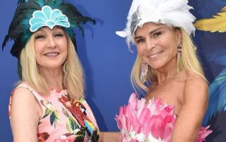 Two women wearing colorful feathered hats and floral dresses against a blue background.