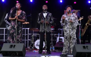 Three vocalists performing on stage with a drum set behind them.