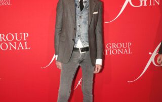 Man posing in a grey suit with vest against a red backdrop.