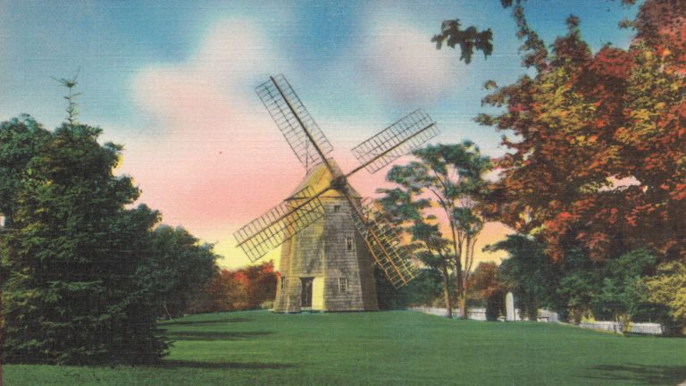 Historic windmill stands amidst green grass and trees under a colorful sky.