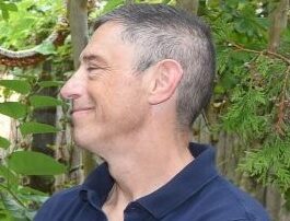 Man in navy shirt smiling outdoors with greenery in the background.