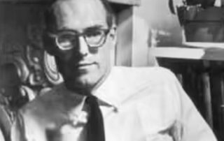 Black and white photo of a man wearing glasses and a tie indoors.