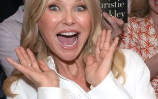 Excited woman with blonde hair smiling widely, hands near her face.