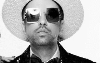 Man wearing a white hat, sunglasses, and a black bow tie.