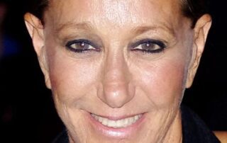 Close-up portrait of a smiling woman with dark eye makeup and slicked-back hair.