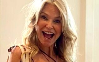 A joyful woman with long blonde hair smiling widely indoors.