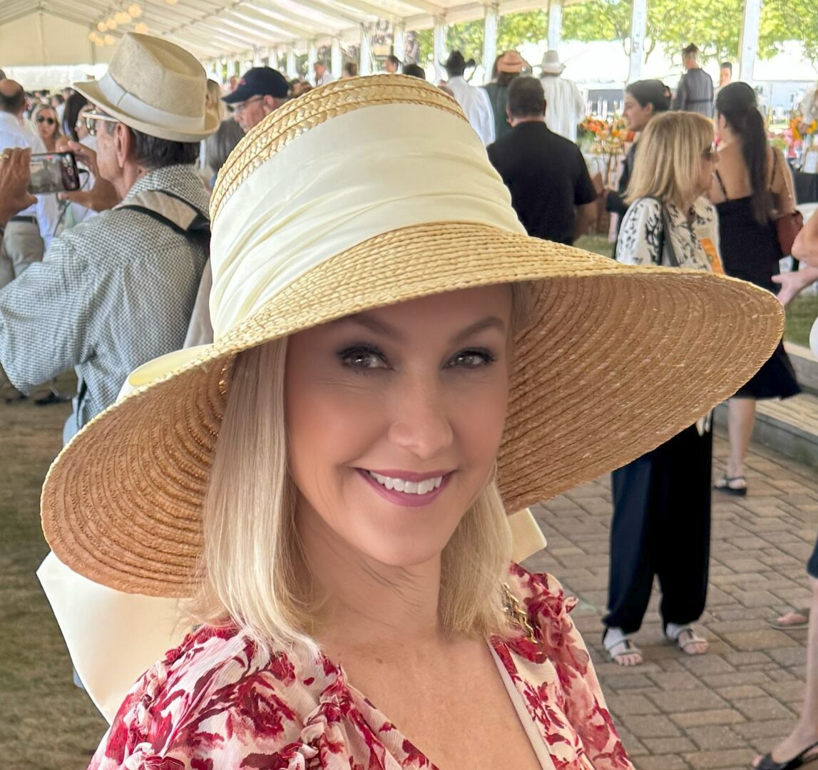 A smiling woman wearing a large straw hat and floral dress at an outdoor event.