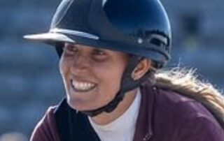 A smiling woman wearing a black equestrian helmet and maroon jacket.