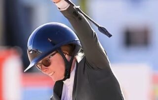 Equestrian rider in helmet and sunglasses raising whip triumphantly.