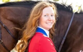 A young woman in a red jacket smiles beside a brown horse.