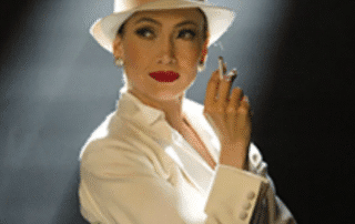 Woman in white suit and hat holding a cigarette, lit dramatically.