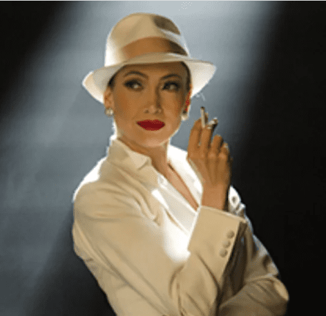 Woman in white suit and hat holding a cigarette, lit dramatically.