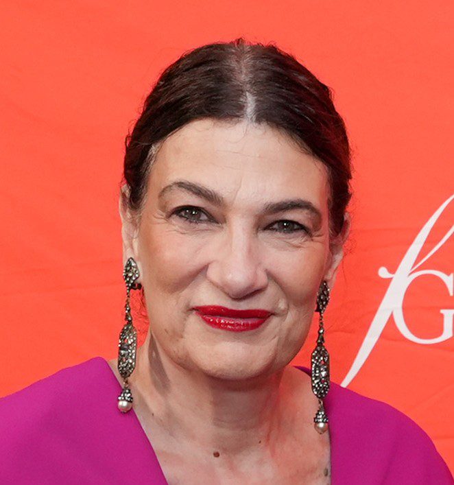 Smiling woman with elegant earrings and bright lipstick against a red backdrop.