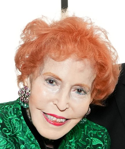 Elderly woman with bright orange hair and vibrant green attire smiling warmly.