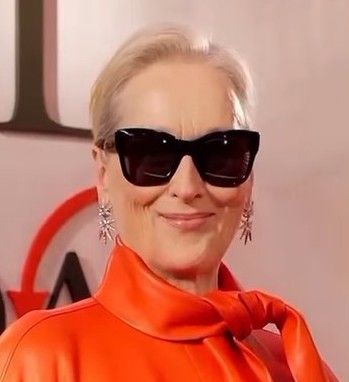 Woman wearing large black sunglasses and an orange outfit, smiling.